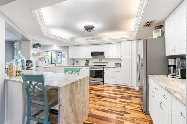 kitchen with white cabinetry, stainless steel appliances, a tray ceiling, a kitchen bar, and light wood-type flooring