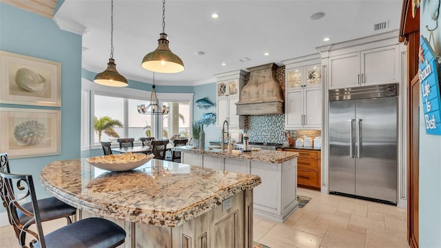 kitchen with stainless steel built in refrigerator, decorative light fixtures, an island with sink, custom exhaust hood, and light stone countertops