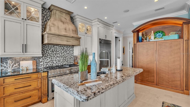 kitchen featuring white cabinets, an island with sink, premium range hood, light stone countertops, and high quality appliances