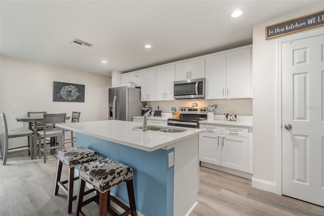 kitchen with white cabinets, stainless steel appliances, an island with sink, sink, and light hardwood / wood-style flooring