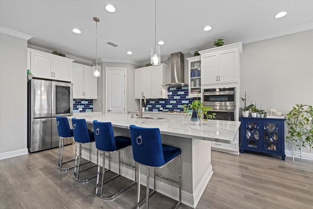 kitchen with a large island with sink, hanging light fixtures, wall chimney exhaust hood, white cabinetry, and stainless steel appliances