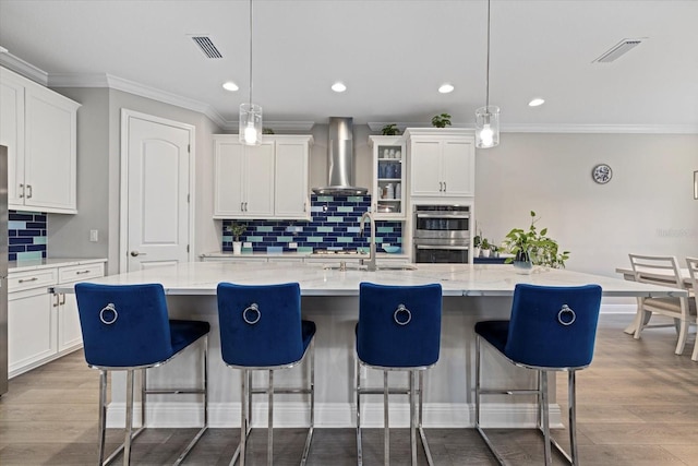 kitchen featuring wall chimney exhaust hood, double oven, an island with sink, pendant lighting, and decorative backsplash