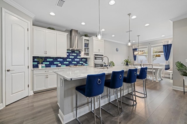 kitchen with white cabinets, pendant lighting, a center island with sink, and wall chimney exhaust hood