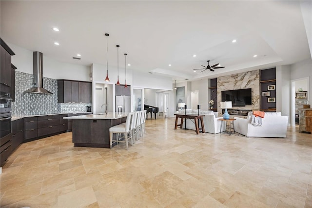 kitchen with decorative light fixtures, an island with sink, decorative backsplash, wall chimney range hood, and a tray ceiling