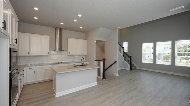 kitchen with oven, white cabinets, wall chimney exhaust hood, an island with sink, and black electric cooktop