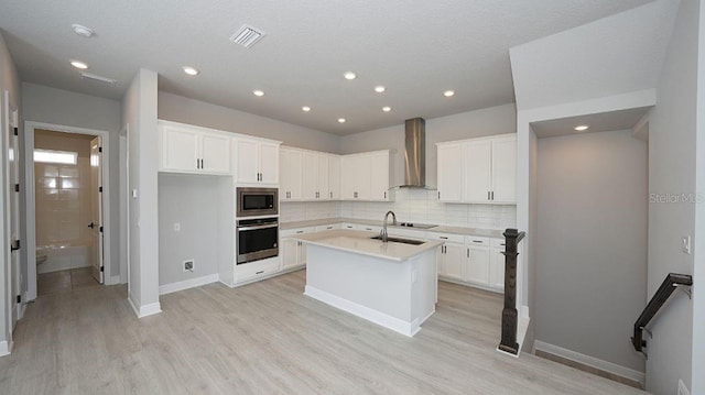 kitchen featuring stainless steel appliances, wall chimney exhaust hood, an island with sink, and white cabinetry