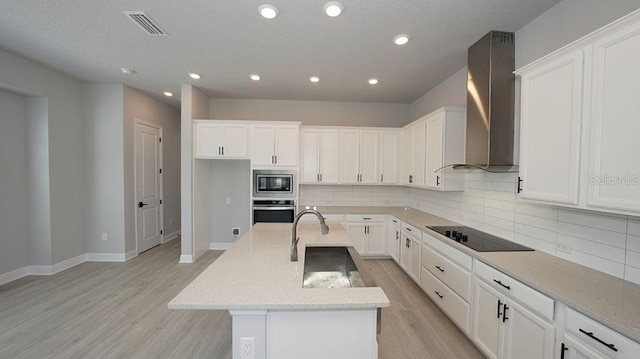 kitchen featuring a kitchen island with sink, stainless steel appliances, light stone counters, wall chimney exhaust hood, and tasteful backsplash