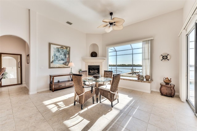 dining area featuring ceiling fan, light tile patterned floors, a tiled fireplace, and a water view