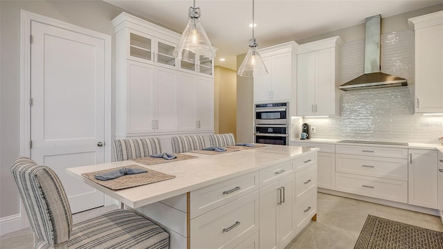 kitchen with white cabinetry, stainless steel double oven, decorative light fixtures, a kitchen breakfast bar, and wall chimney range hood