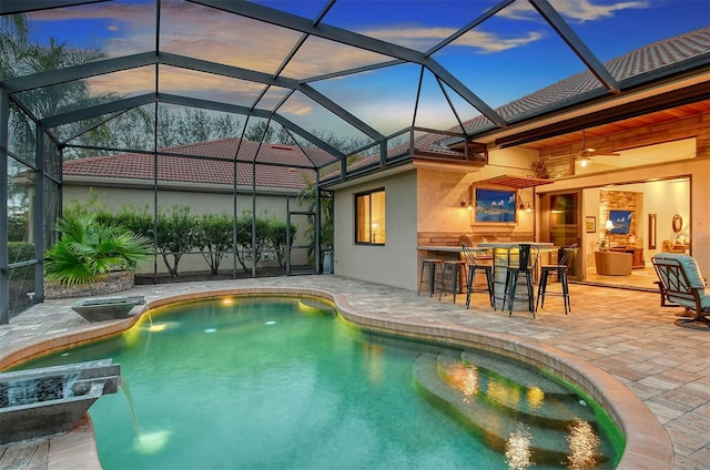 pool at dusk featuring ceiling fan, a bar, a patio, and glass enclosure