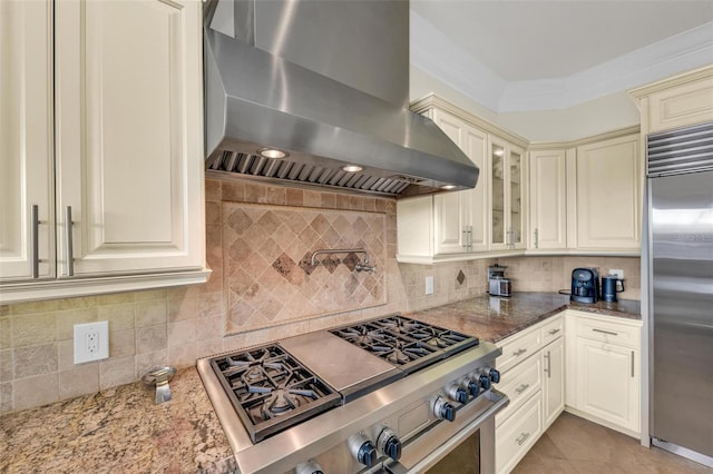 kitchen with range hood, decorative backsplash, built in refrigerator, dark stone counters, and cream cabinetry