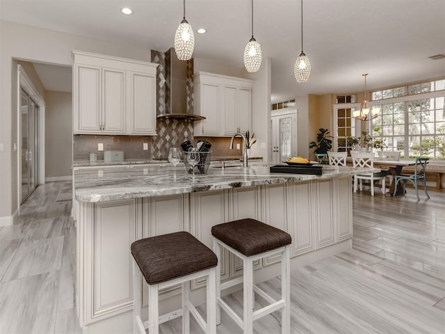 kitchen featuring wall chimney range hood, white cabinetry, hanging light fixtures, and light stone counters