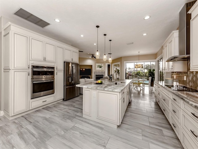 kitchen with wall chimney range hood, decorative light fixtures, stainless steel appliances, light stone counters, and a center island with sink