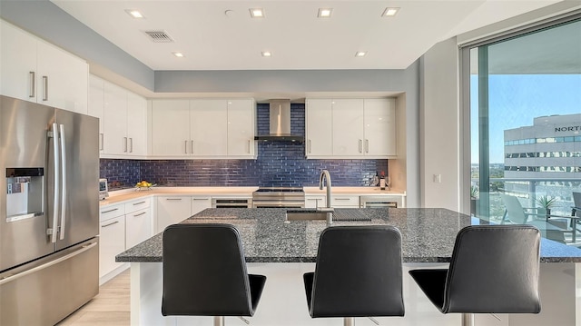 kitchen featuring white cabinetry, wall chimney exhaust hood, a kitchen bar, a center island with sink, and appliances with stainless steel finishes