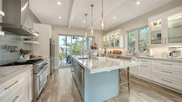 kitchen featuring wall chimney range hood, beam ceiling, premium appliances, an island with sink, and white cabinets