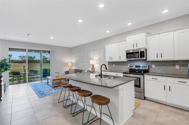 kitchen with white cabinetry, stainless steel appliances, dark stone countertops, sink, and a kitchen island with sink