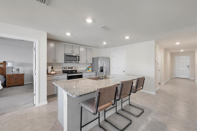 kitchen with gray cabinets, appliances with stainless steel finishes, an island with sink, light stone counters, and sink