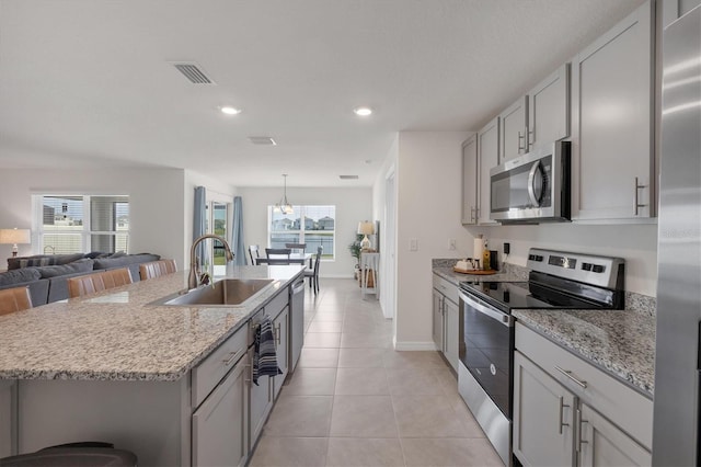 kitchen with appliances with stainless steel finishes, an island with sink, light tile patterned floors, light stone counters, and sink