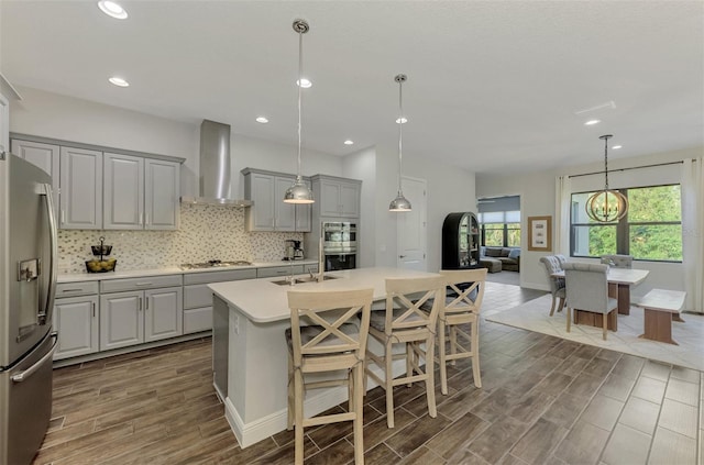 kitchen featuring wall chimney exhaust hood, pendant lighting, a kitchen island with sink, gray cabinetry, and appliances with stainless steel finishes