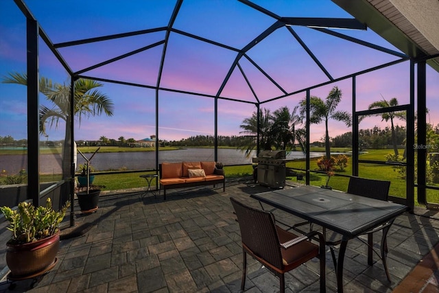 patio terrace at dusk with a lanai, outdoor lounge area, a grill, a lawn, and a water view