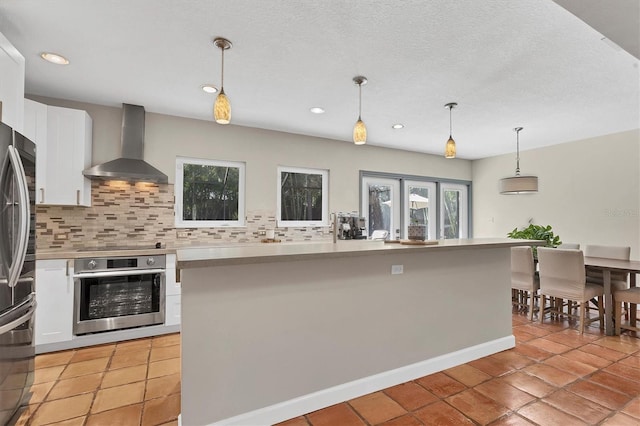 kitchen with stainless steel appliances, white cabinets, decorative backsplash, hanging light fixtures, and wall chimney range hood