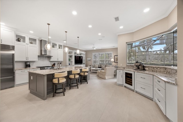 kitchen with built in fridge, an island with sink, white cabinets, and wall chimney exhaust hood