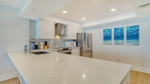 kitchen with kitchen peninsula, stainless steel appliances, wall chimney range hood, white cabinetry, and sink