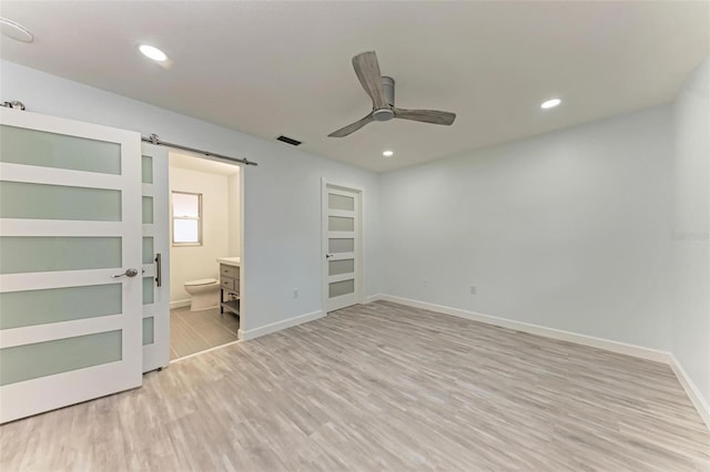 unfurnished bedroom with ensuite bathroom, ceiling fan, light hardwood / wood-style floors, and a barn door