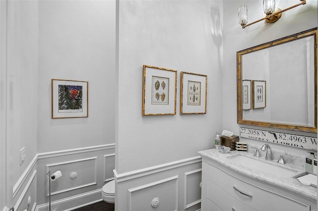 half bath with vanity, a decorative wall, toilet, and wainscoting