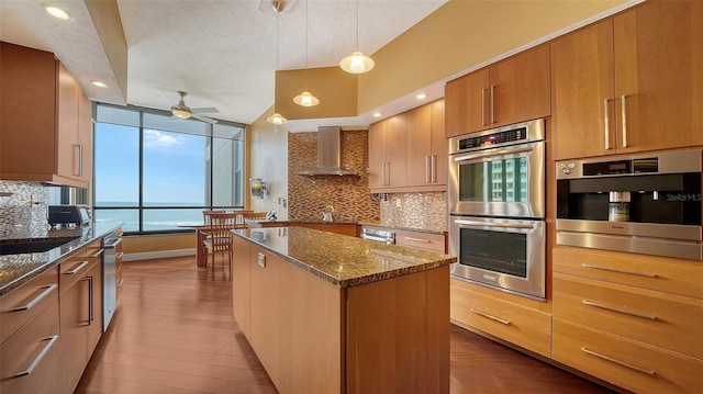 kitchen with wall chimney exhaust hood, pendant lighting, a center island, stainless steel double oven, and a water view