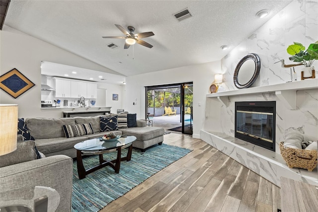 living room with vaulted ceiling, a textured ceiling, light wood-type flooring, ceiling fan, and a high end fireplace