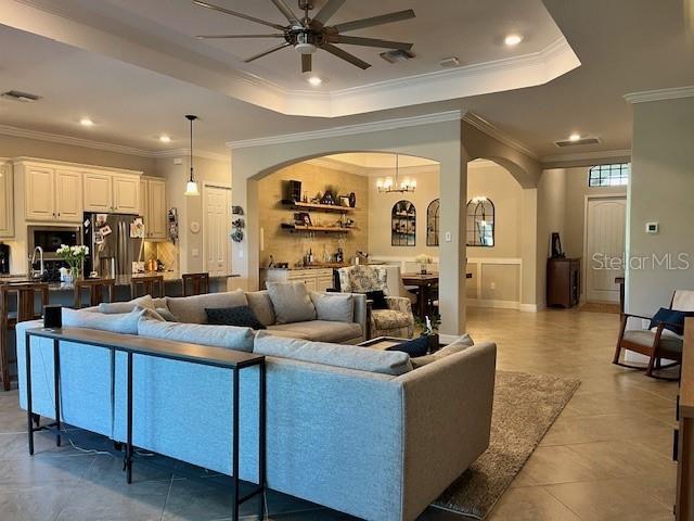 living room with a raised ceiling, tile patterned floors, ceiling fan with notable chandelier, and crown molding