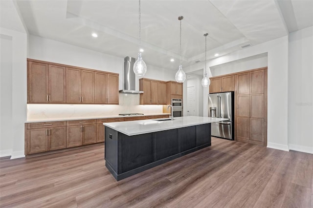 kitchen featuring a center island with sink, stainless steel appliances, a tray ceiling, decorative light fixtures, and wall chimney range hood