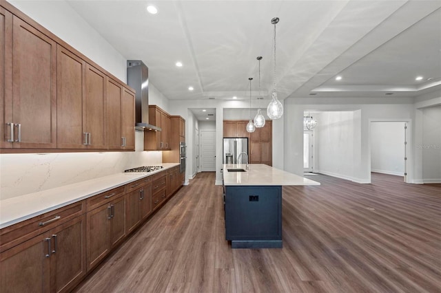 kitchen featuring appliances with stainless steel finishes, a kitchen island with sink, dark wood-type flooring, hanging light fixtures, and wall chimney range hood