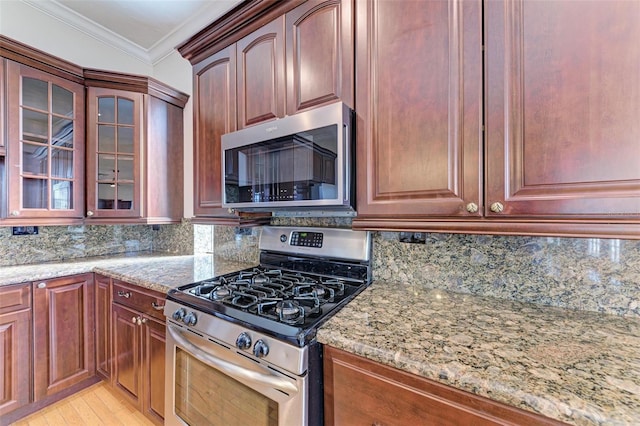 kitchen with appliances with stainless steel finishes, glass insert cabinets, light stone countertops, and crown molding