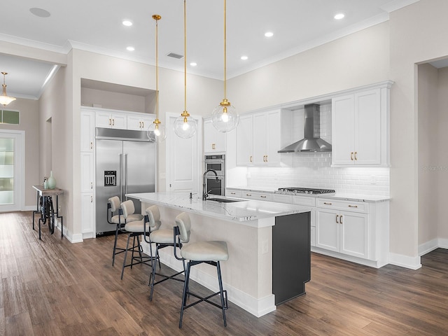 kitchen with appliances with stainless steel finishes, wall chimney exhaust hood, a center island with sink, and pendant lighting