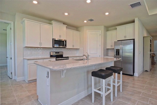 kitchen featuring an island with sink, stainless steel appliances, light stone counters, white cabinets, and a breakfast bar