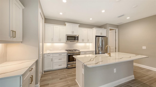 kitchen featuring stainless steel appliances, white cabinetry, light stone counters, and an island with sink