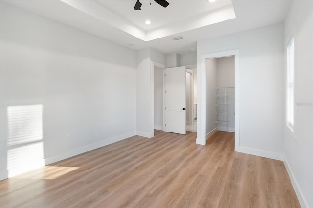 unfurnished bedroom with a walk in closet, a closet, ceiling fan, a tray ceiling, and light hardwood / wood-style flooring