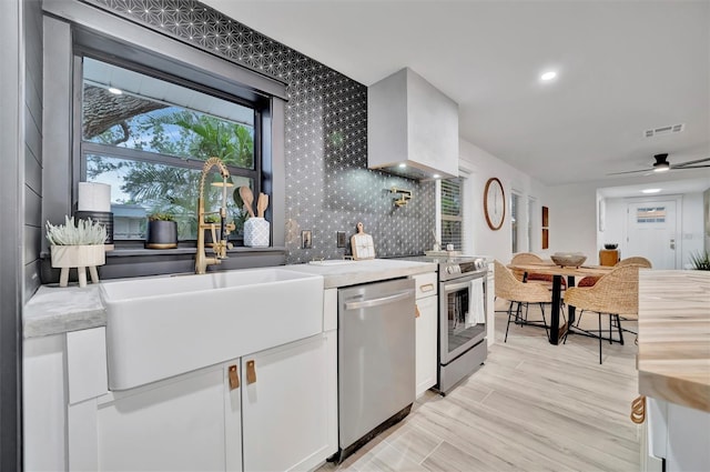 kitchen with stainless steel appliances, ceiling fan, wall chimney exhaust hood, sink, and white cabinetry