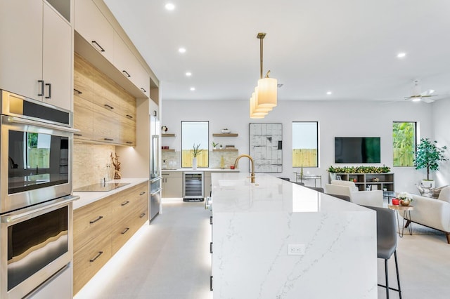 kitchen featuring wine cooler, an island with sink, black electric stovetop, double oven, and decorative light fixtures