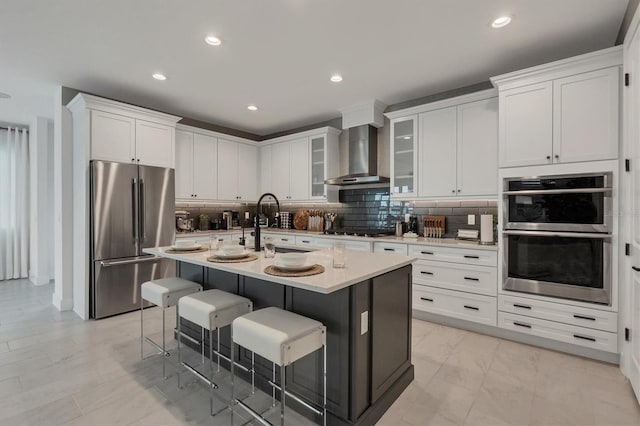 kitchen featuring wall chimney range hood, a center island with sink, white cabinetry, appliances with stainless steel finishes, and a breakfast bar area