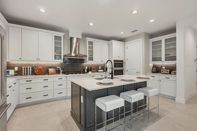 kitchen with wall chimney range hood, sink, white cabinetry, a kitchen island with sink, and a breakfast bar area