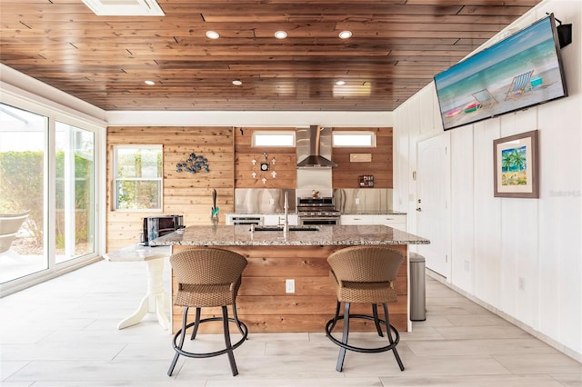 kitchen with wall chimney range hood, stone counters, stainless steel range, wood ceiling, and a breakfast bar area