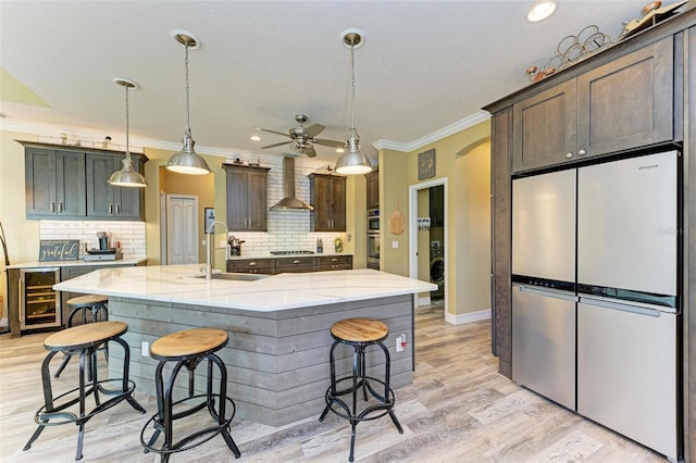 kitchen with beverage cooler, wall chimney exhaust hood, stainless steel appliances, sink, and a kitchen island with sink