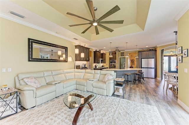 living room with ceiling fan, a tray ceiling, crown molding, and light wood-type flooring