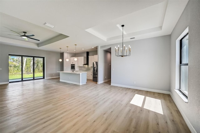 unfurnished living room featuring a raised ceiling, ceiling fan with notable chandelier, and light wood-type flooring