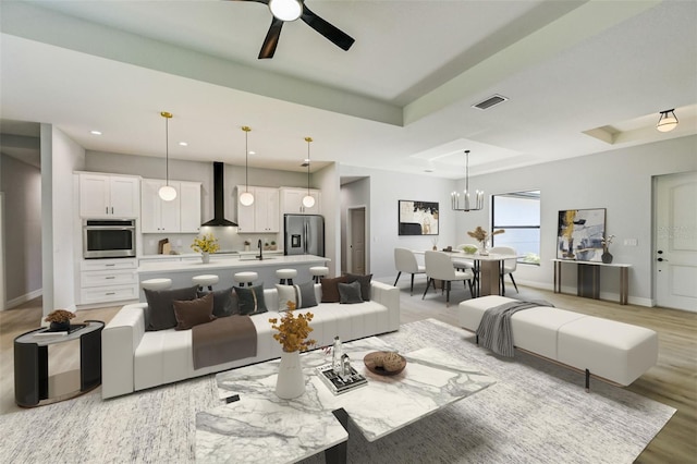 living room featuring ceiling fan with notable chandelier, a tray ceiling, sink, and light wood-type flooring