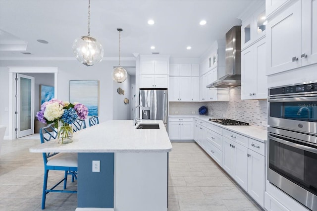 kitchen featuring white cabinets, wall chimney exhaust hood, decorative light fixtures, stainless steel appliances, and a kitchen island with sink