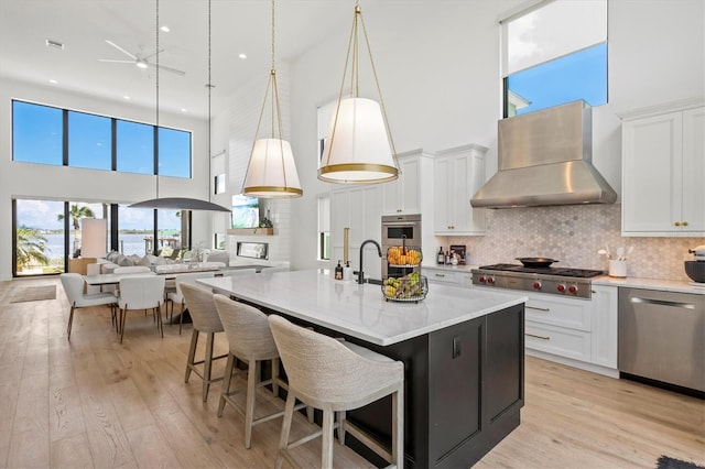 kitchen with decorative light fixtures, wall chimney range hood, stainless steel appliances, a kitchen island with sink, and white cabinets